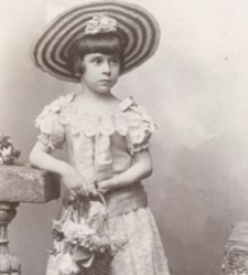 Dziewczynka w koronkowej sukience, kapeluszu, w rękach trzyma koszyczek z kwiatami, oparta o balustradę