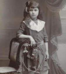 Dziewczynka w sukience z białym , koronkowym kołnierzem , w ręku trzyma kółko , oparta o fotel