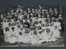 Komunia święta, dziewczynki w białych sukienkach, chłopcy przeważnie w ciemnych ubrankach z białymi kołnierzykami pomiędzy dziećmi siedzi ksiądz i dwie kobiety