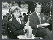 Kobieta w harcerskim mundurze kroi bochen chleba, obok niej siedzi I sekretarz Komitetu Wojewódzkiego PZPR w Białymstoku Zdzisław Kurowski z harcerzami