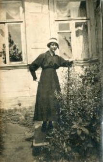 Kobieta w ciemnej sukni i białym kapeluszu przy roślinach, za nią drewniany dom