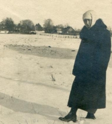 Kobieta w długim płaszczu i w białej chustce na głowie, za nią zabudowania i pola