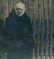 Kobieta w ciemnym, zimowym płaszczu i białej chustce na tle drewnianego domu