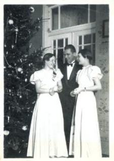 Dwie kobiety w jasnych sukniach i mężczyzna w garniturze przy choince
