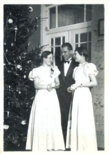 Dwie kobiety w jasnych sukniach i mężczyzna w garniturze przy choince